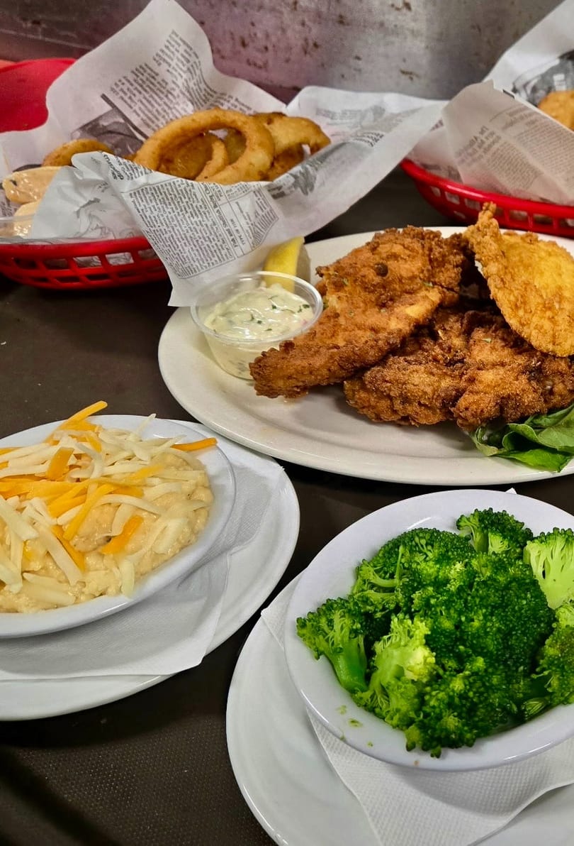Fried fish, onion rings, grits with cheese, and steamed broccoli on a dark table.
