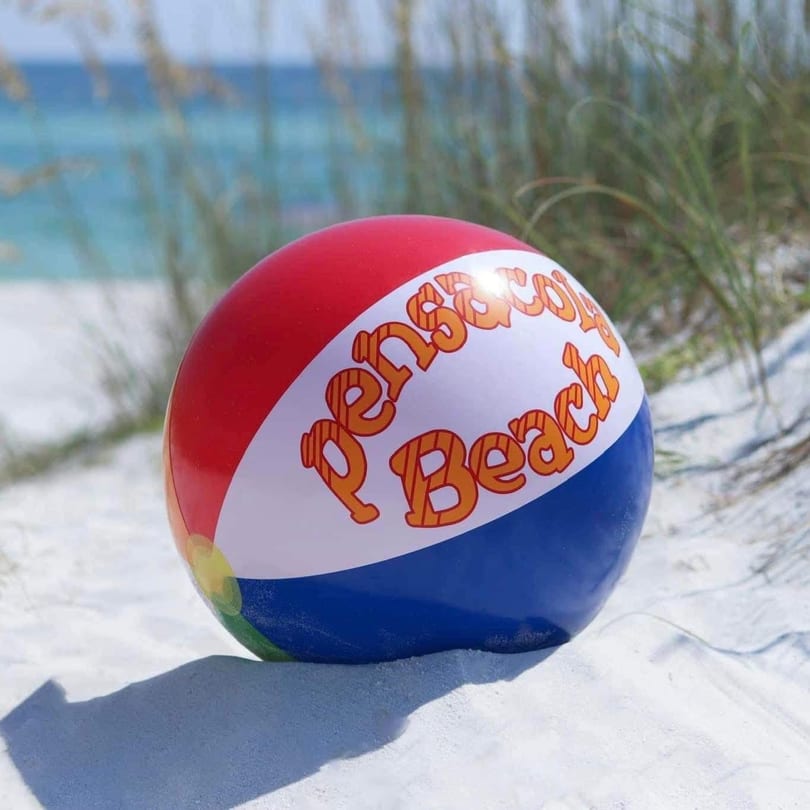 A red, white, and blue Pensacola Beach ball sits on white sand with the ocean in the background.