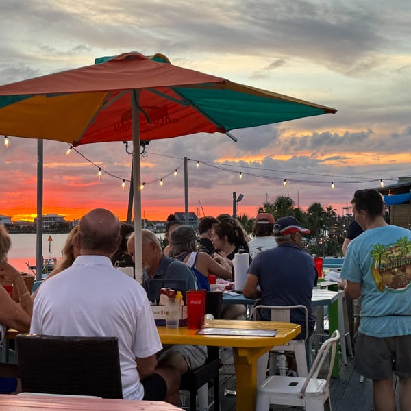 Diners on an outdoor patio at sunset, with a vibrant orange sky over the water.