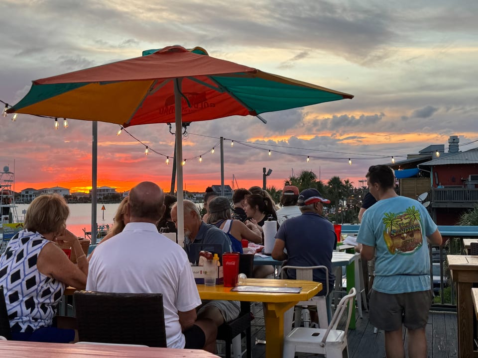Diners on an outdoor patio at sunset, with a vibrant orange sky over the water.