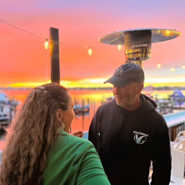 Two people on a restaurant patio at sunset, with boats and water in the background.
