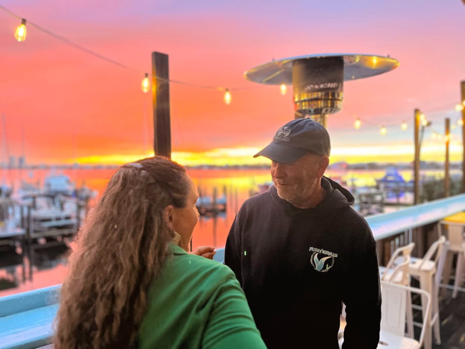 Two people on a restaurant patio at sunset, with boats and water in the background.