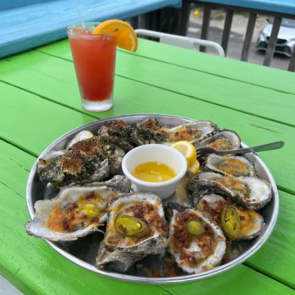 A platter of baked oysters with various toppings, a cocktail, on a vibrant green table by the water.