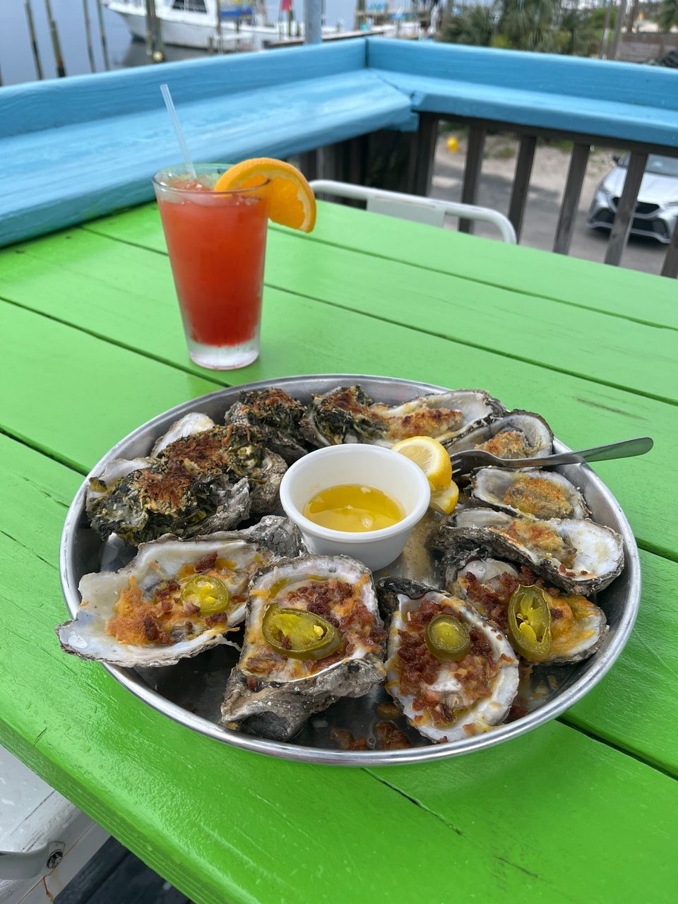 A platter of baked oysters with various toppings, a cocktail, on a vibrant green table by the water.