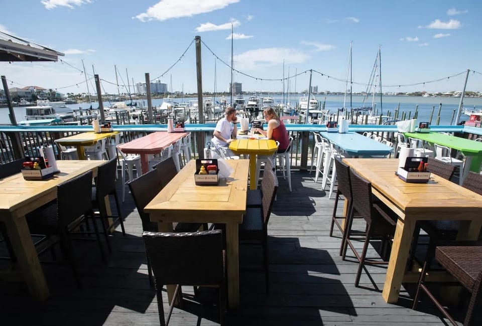 Diners on a sunny outdoor patio overlooking a marina with boats and distant buildings.