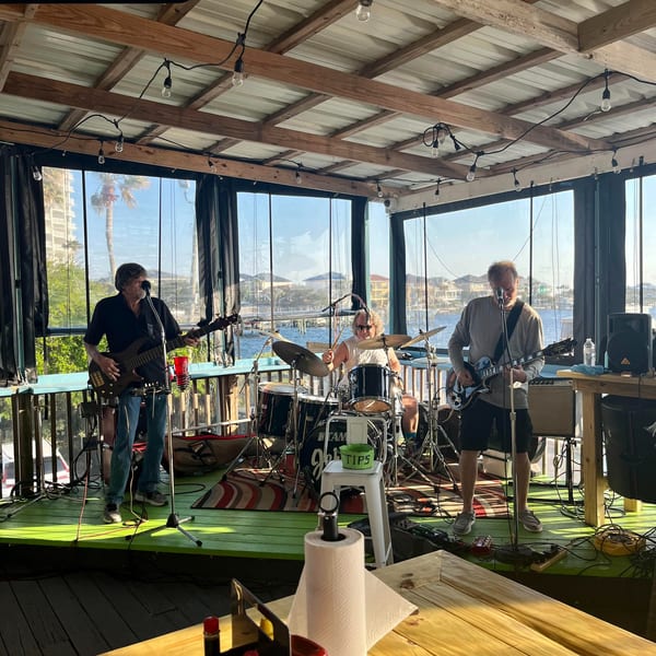 A band performs on a green stage overlooking a sunny waterfront with buildings and palm trees.