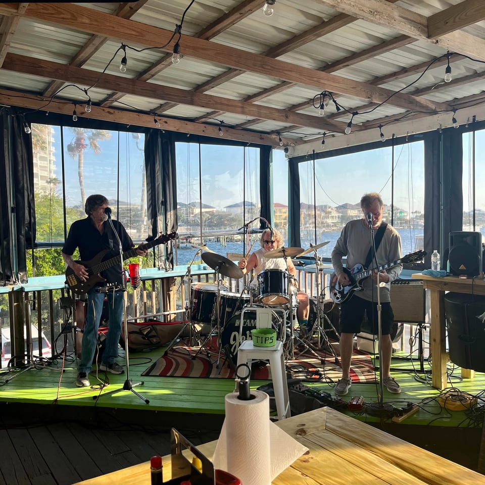 A band performs on a green stage overlooking a sunny waterfront with buildings and palm trees.