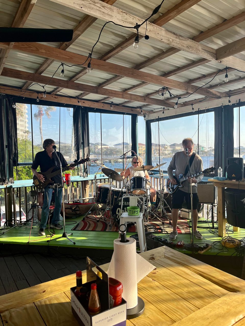 A band performs on a green stage overlooking a sunny waterfront with buildings and palm trees.