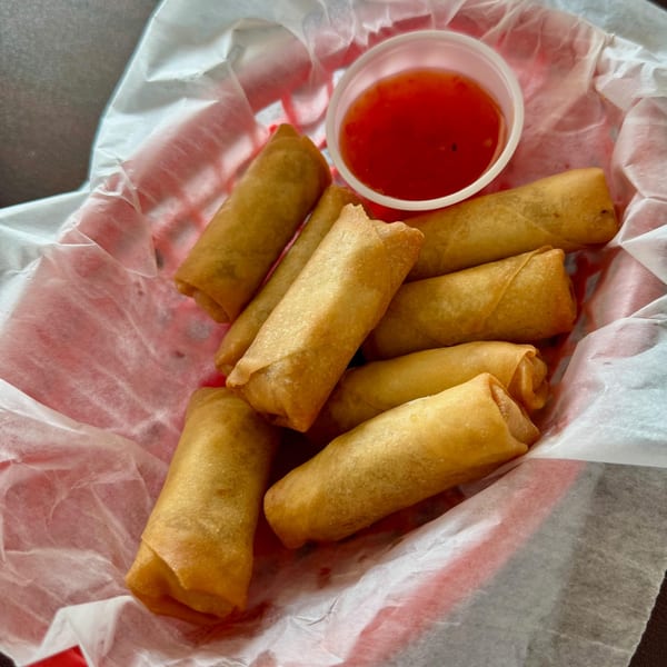 Golden-brown spring rolls and a small cup of red dipping sauce in a red basket lined with white paper.