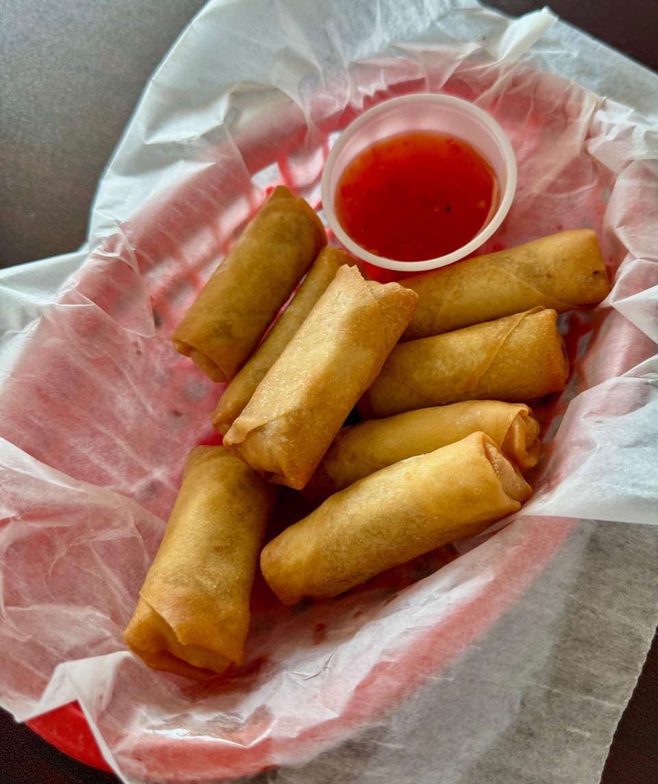 Golden-brown spring rolls and a small cup of red dipping sauce in a red basket lined with white paper.